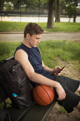 Mann mit Basketball, der sein Smartphone benutzt, während er im Park auf einer Bank sitzt - CAVF48454