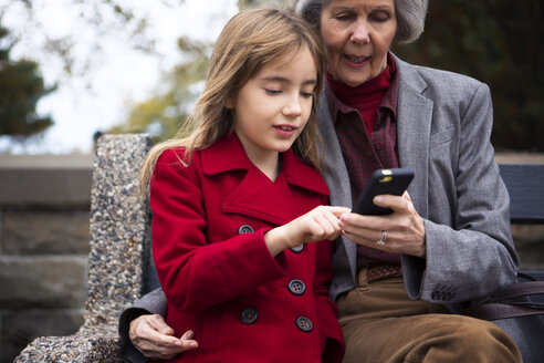 Familie benutzt Mobiltelefon, während sie auf einer Bank im Park sitzt - CAVF48563