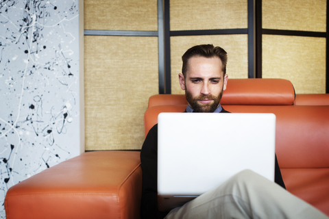 Mann, der einen Laptop benutzt, während er auf dem Sofa im Büro sitzt, lizenzfreies Stockfoto