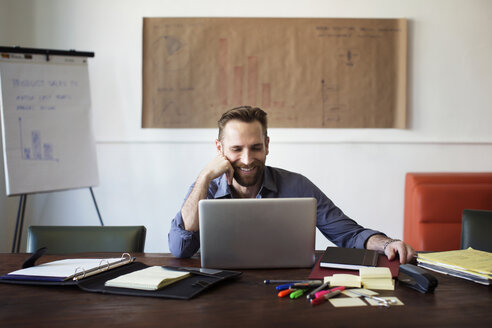 Smiling businessman looking at laptop computer while sitting in office - CAVF48643