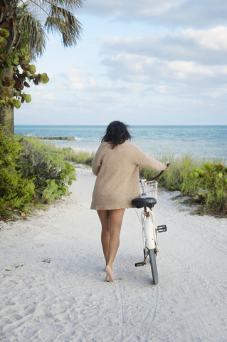 Rückansicht einer Frau mit Fahrrad, die auf Sand gegen das Meer läuft, lizenzfreies Stockfoto