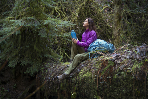 Frau hält Flasche und schaut nach oben, während sie auf einem umgestürzten Baumstamm im Wald sitzt, lizenzfreies Stockfoto