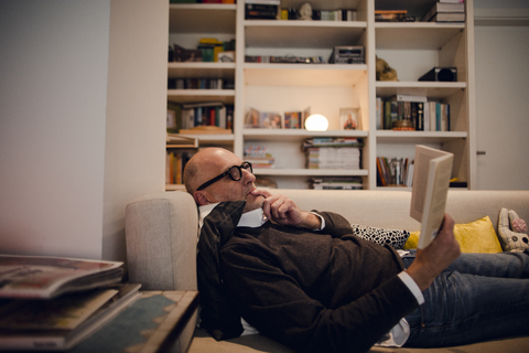 Älterer Mann liegt auf der Couch und liest ein Buch, lizenzfreies Stockfoto