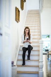 Smiling woman sitting on stairs at home using cell phone - MOEF01103