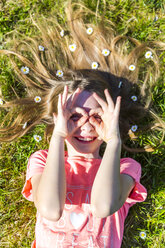 Lächelndes Mädchen im Gras liegend im Frühling mit Gänseblümchen auf dem Haar, das mit den Fingern eine Brille formt - SARF03680