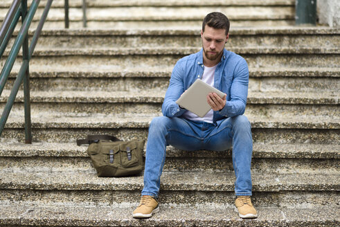 Junger Mann sitzt auf einer Treppe, arbeitet und benutzt ein digitales Tablet - JSMF00144