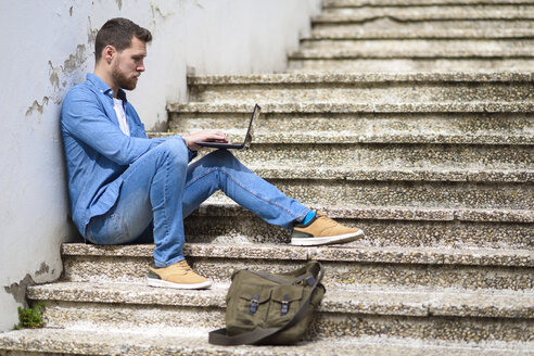 Junger Mann sitzt auf einer Treppe, arbeitet, benutzt einen Laptop - JSMF00145