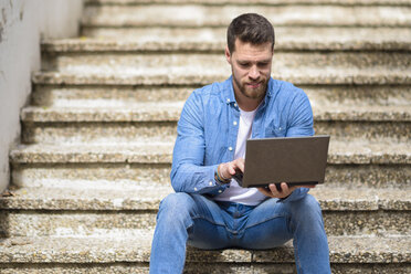 Junger Mann sitzt auf einer Treppe, arbeitet, benutzt einen Laptop - JSMF00146