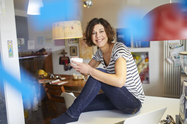 Porträt einer glücklichen reifen Frau, die mit einer Tasse Kaffee am Küchentisch sitzt - PNEF00620
