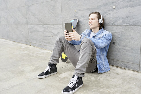 Junger Mann mit Skateboard auf dem Boden sitzend, mit digitalem Tablet - FMOF00367