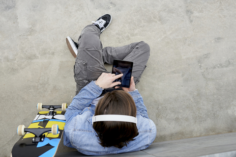 Junger Mann mit Skateboard auf dem Boden sitzend, mit digitalem Tablet, lizenzfreies Stockfoto