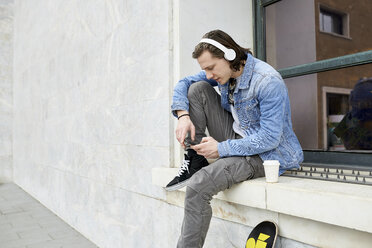 Young man sitting on window sill, waiting, using smartphone - FMOF00374