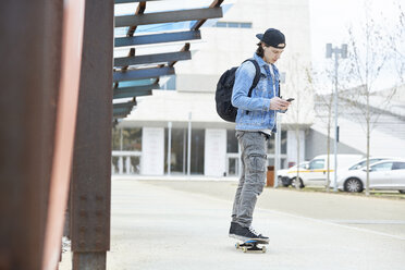 Junger Mann, der in der Stadt Skateboard fährt, sein Smartphone benutzt und Musik hört - FMOF00379