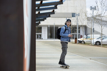 Junger Mann, der in der Stadt Skateboard fährt, sein Smartphone benutzt und Musik hört - FMOF00380