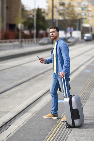 Junger Mann wartet auf einem Bahnhof mit Smartphone in der Hand und Einkaufswagen, lizenzfreies Stockfoto