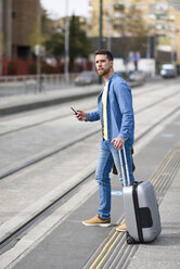Young man waiting at a station with smartphone in his hand and trolley - JSMF00154