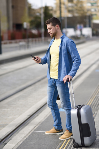 Junger Mann wartet auf einem Bahnhof mit Smartphone in der Hand und Einkaufswagen, lizenzfreies Stockfoto