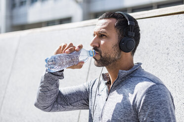 Mann, der eine Pause vom Sport macht, Kopfhörer trägt und aus einer Flasche trinkt - DIGF04057