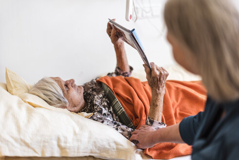 Frau kümmert sich um eine alte Frau, die im Bett liegt und ein Buch liest - DIGF04065