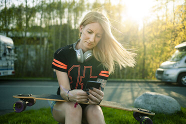 Porträt einer blonden Frau mit Longboard, die am Straßenrand sitzt und ihr Smartphone benutzt - NAF00088