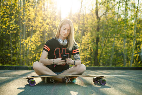 Blonde Frau mit Longboard sitzt auf der Straße und benutzt ihr Smartphone, lizenzfreies Stockfoto