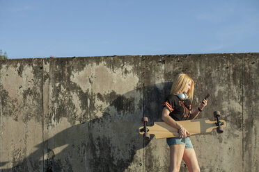 Blond woman with longboard standing in front of wall looking at smartphone - NAF00091