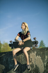 Blonde Frau mit Longboard an der Wand sitzend im Sonnenlicht - NAF00092