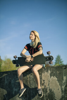 Blond woman with longboard sitting on wall at sunlight - NAF00092