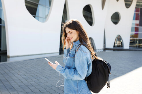 Spanien, Barcelona, lächelnde junge Frau mit Rucksack hört Musik mit Handy und Kopfhörer - VABF01554