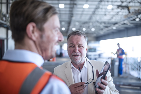 Businessman showing cell phone to man in reflective vest in industrial hall stock photo