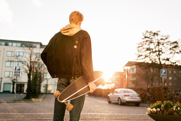 Rear view of young man holding skateboard while standing on sidewalk in city - MASF07087