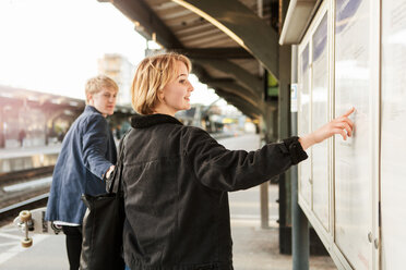 Junger Mann, der ein Teenager-Mädchen zieht, das auf eine Informationstafel am Bahnsteig zeigt - MASF07094