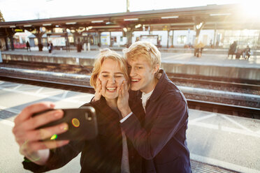 Fröhliches junges Paar nimmt Selfie durch Handy auf Bahnhofsplattform - MASF07096