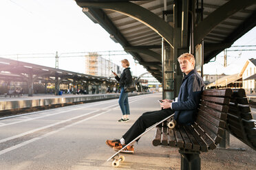 Junger Mann sitzt auf einer Bank mit einem Mädchen im Hintergrund am Bahnsteig - MASF07097