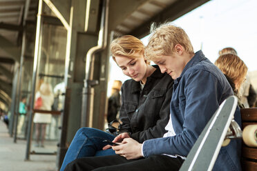 Junger Mann zeigt Teenager-Mädchen Handy, während er auf dem Bahnsteig sitzt - MASF07100