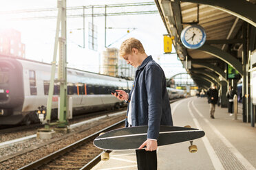 Side view of young man using mobile phone while holding skateboard on railroad station platform - MASF07104