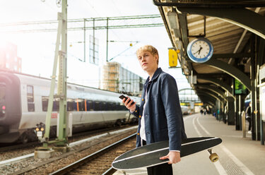 Young man holding mobile phone and skateboard while waiting on railroad station platform - MASF07105