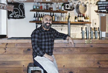 Confident smiling bartender sitting by bar counter - MASF07127