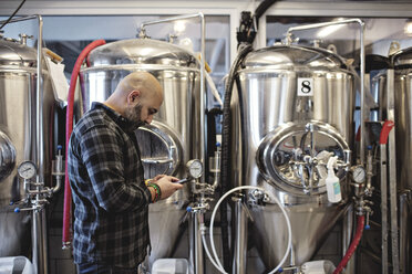 Seitenansicht eines Managers, der ein Mobiltelefon benutzt, während er bei den Lagertanks einer Brauerei steht - MASF07170