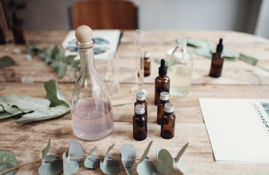 Blick von oben auf verschiedene Parfümflaschen inmitten von Blättern und Broschüren auf einem Tisch in einer Werkstatt - MASF07191