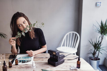 Woman smelling leaves while sitting at table against wall in perfume workshop - MASF07201