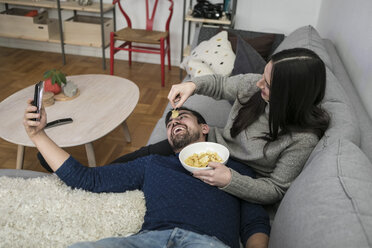 Happy man taking selfie while woman feeding chips to him on sofa - MASF07257