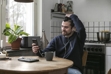 Mann mit Kopfhörern, der ein Mobiltelefon benutzt, während er am Tisch in der Küche sitzt - MASF07258