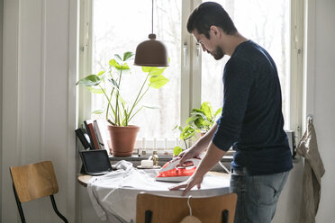 Seitenansicht eines Mannes, der ein Hemd auf einem Tisch am Fenster bügelt, während er in der Küche steht - MASF07267