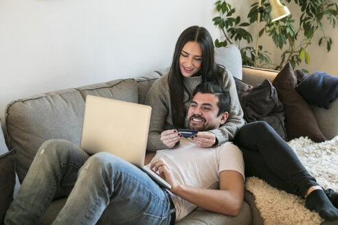Ein glückliches Paar benutzt seine Kreditkarte beim Online-Einkauf mit dem Laptop auf dem Sofa zu Hause - MASF07285