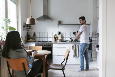 Smiling man washing utensils while woman having drink at table in kitchen - MASF07291