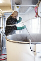 Männlicher Arbeiter rührt bei der Arbeit in einer Brauerei Bier in einem Behälter um - MASF07295