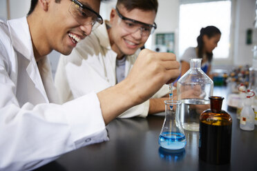 Smiling young male students mixing solutions in glassware at chemistry laboratory - MASF07308