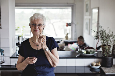 Confident senior woman holding smart phone while listening to in-ear headphones in kitchen at home - MASF07389