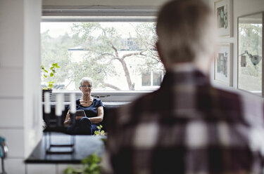 Rückansicht eines älteren Mannes mit Blick auf eine Frau, die auf dem Sofa im Wohnzimmer sitzt - MASF07404